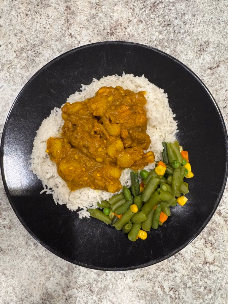 Jamaican curry chicken on a bed of white rice next to steamed mixed vegetables all on a black plate