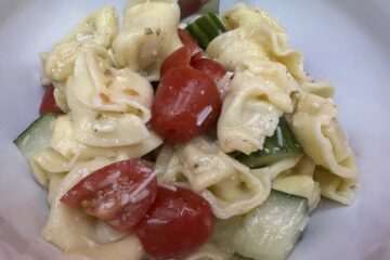 Tortellini cucumbers and tomatoes with pieces of Parmesan cheese and Italian dressing all mixed together in a white bowl