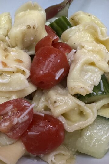 Tortellini cucumbers and tomatoes with pieces of Parmesan cheese and Italian dressing all mixed together in a white bowl