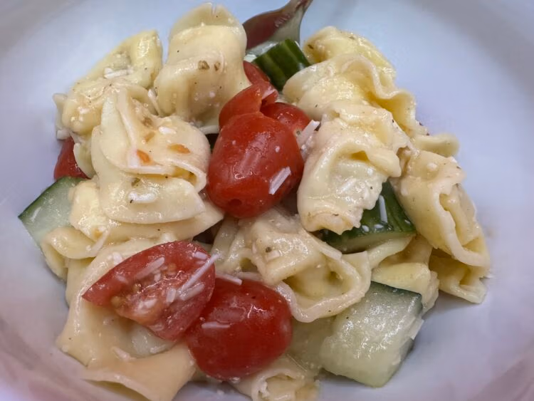 Tortellini cucumbers and tomatoes with pieces of Parmesan cheese and Italian dressing all mixed together in a white bowl