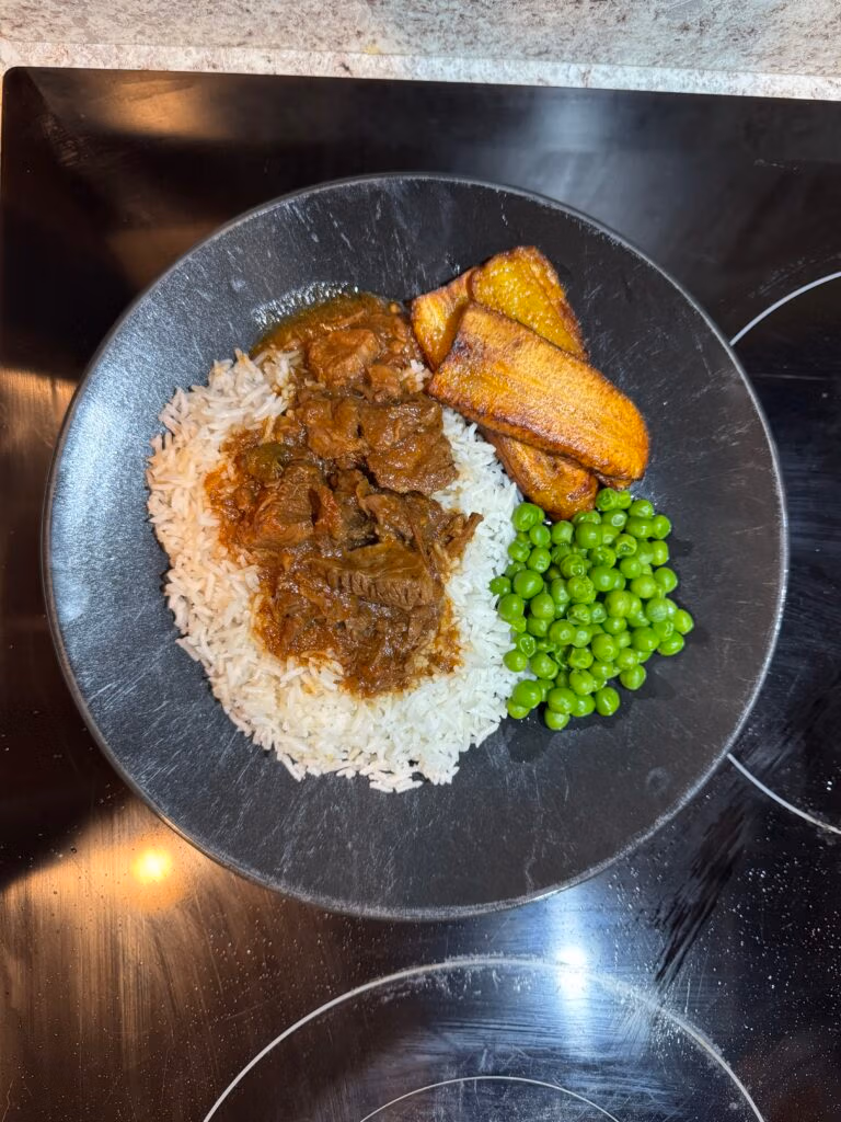 Jamaican brown stewed beef served over a bed of white rice next to fried plantains and steamed sweet peas all on a black plate