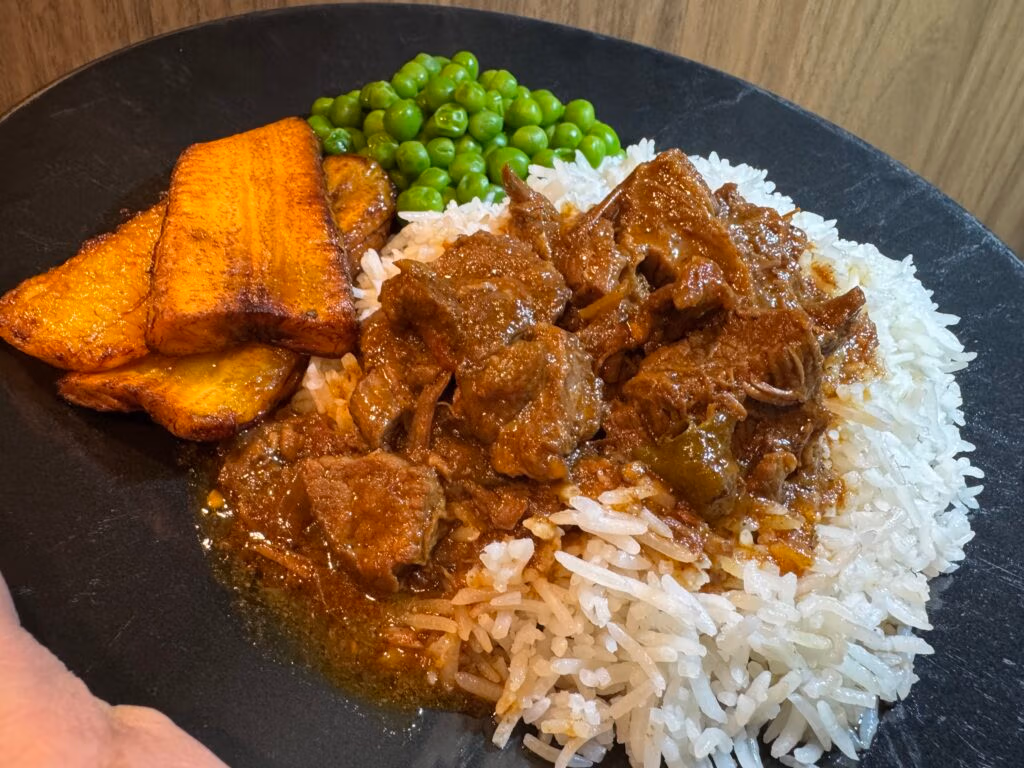 Jamaican brown stewed beef served over a bed of white rice next to fried plantains and steamed sweet peas all on a black plate