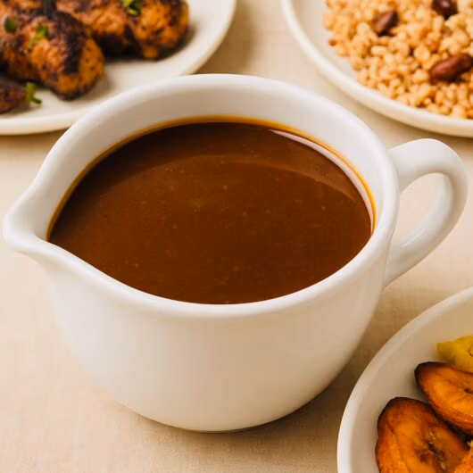 A white gravy boat filled with rich, glossy jerk sauce, surrounded by Jamaican side dishes including rice and peas, fried plantains, and grilled jerk chicken, captured with soft natural lighting on a beige tablecloth.
