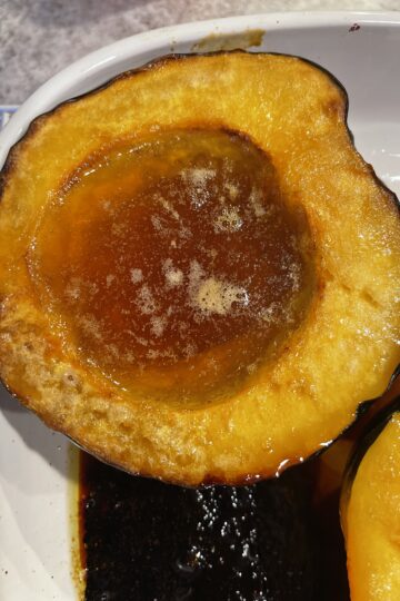 Two halves of golden roasted acorn squash with bubbling maple syrup and butter, caramelized edges, and a fork resting beside them.