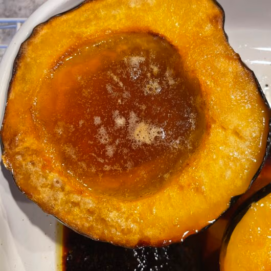 Two halves of golden roasted acorn squash with bubbling maple syrup and butter, caramelized edges, and a fork resting beside them.