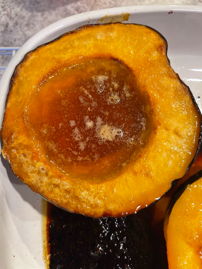 Two halves of golden roasted acorn squash with bubbling maple syrup and butter, caramelized edges, and a fork resting beside them.