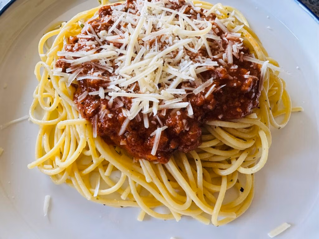 A white plate of cooked spaghetti with a beef pasta sauce and parmesan garnish
