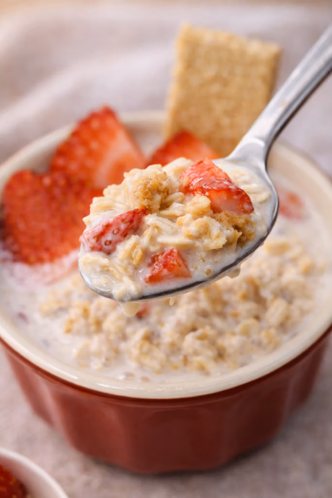 A small red bowl with strawberries and grahm cracker of overnight oats with a spoonful of strawberry shortcake overnight oats